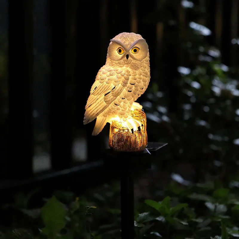 Fehér bagolylámpa sárga szemekkel, melegen világító fatörzs alakú talapzaton, sötét kerti háttér előtt. 6