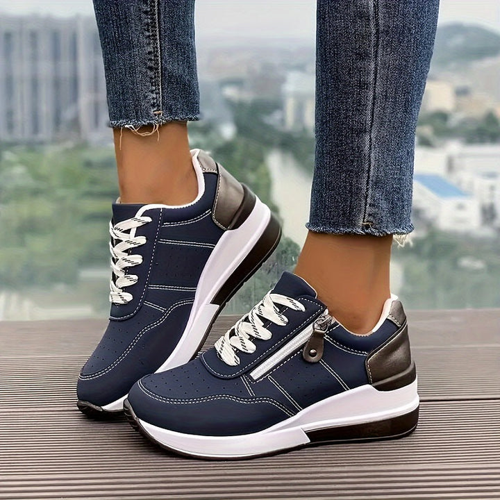 Sötétkék, fűzős, oldalcipzáras sneaker farmeres lábon, városi kilátás előtt, vastag fehér talppal 1