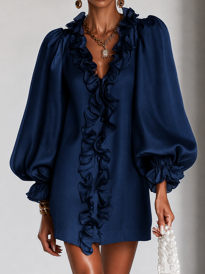 Sötétkék ruha közeli elölnézetben, a fodros V-nyakkivágás és a puffos ujjak részletével, arany nyaklánccal. 6