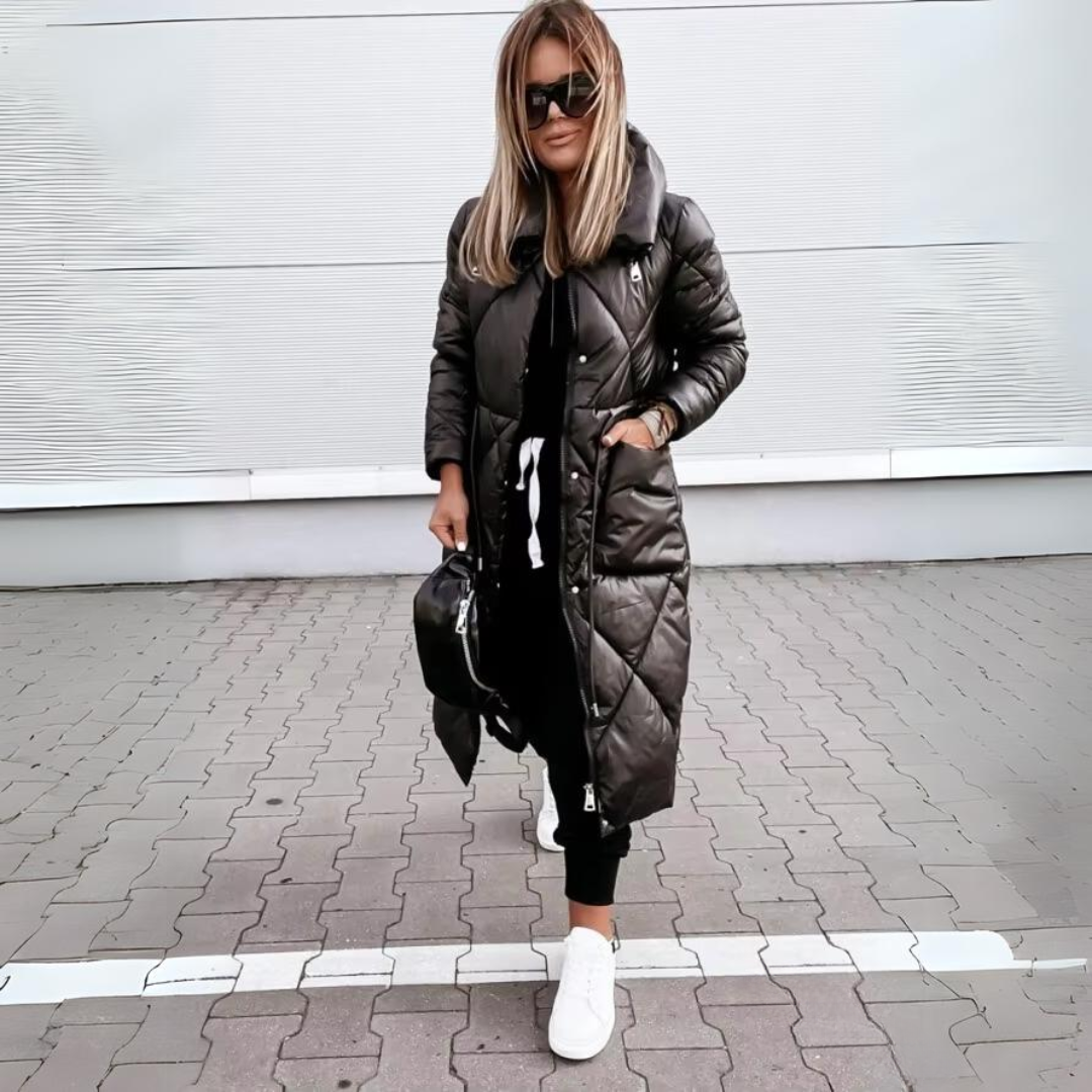 Modell elölről, becipzározott fekete hosszú steppelt kabátban, zsebében kézzel, fekete nadrággal, fehér sportcipővel, térkövezett úton állva. 5