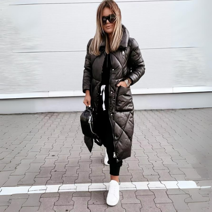 Modell elölről, becipzározott fekete hosszú steppelt kabátban, zsebében kézzel, fekete nadrággal, fehér sportcipővel, térkövezett úton állva. 5