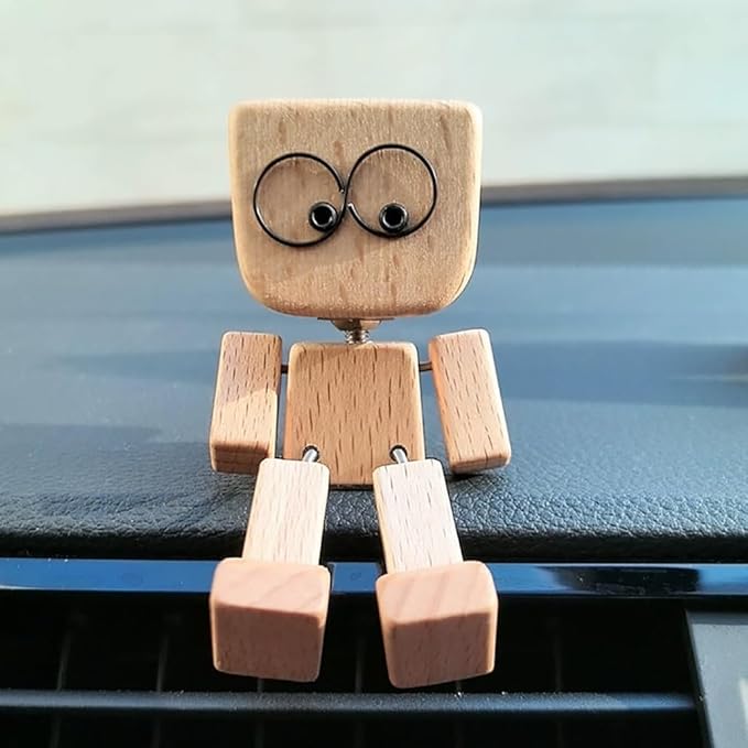 Szemből látható, világos fából készült ülő figura nagy, kerek szemű arccal egy autó műszerfalán, lába lelóg a szellőzőrácsra. 1