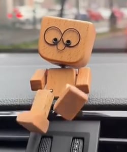 Közeli kép a nagy, kerek szemű arcú fa figuráról, amint egy autó műszerfalának szélén ül, lábát lógatva. 6