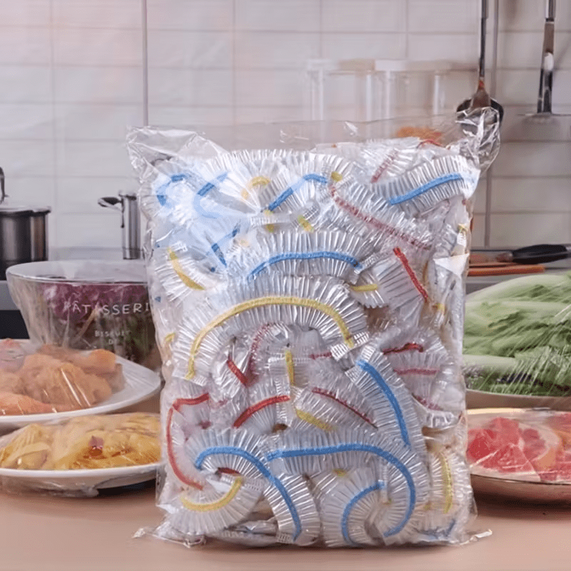 Átlátszó zacskónyi, színes gumiszalagos eldobható fedő egy konyhapulton, mellette fóliával letakart ételek tányérokon. 1