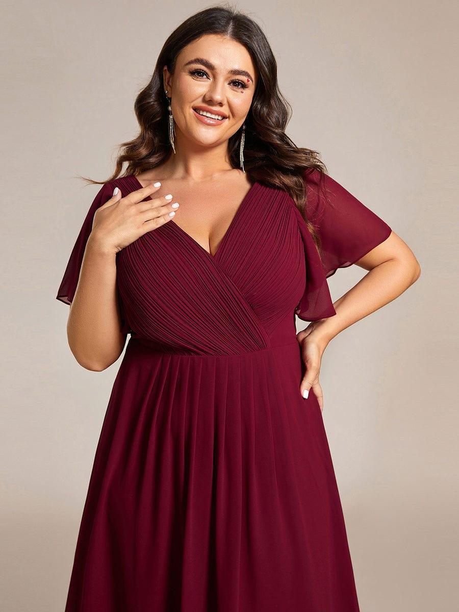 Közelkép a bordó ruha redőzött mellrészéről és fodros ujjairól, a plus size modell kezét a mellkasához emeli. 51