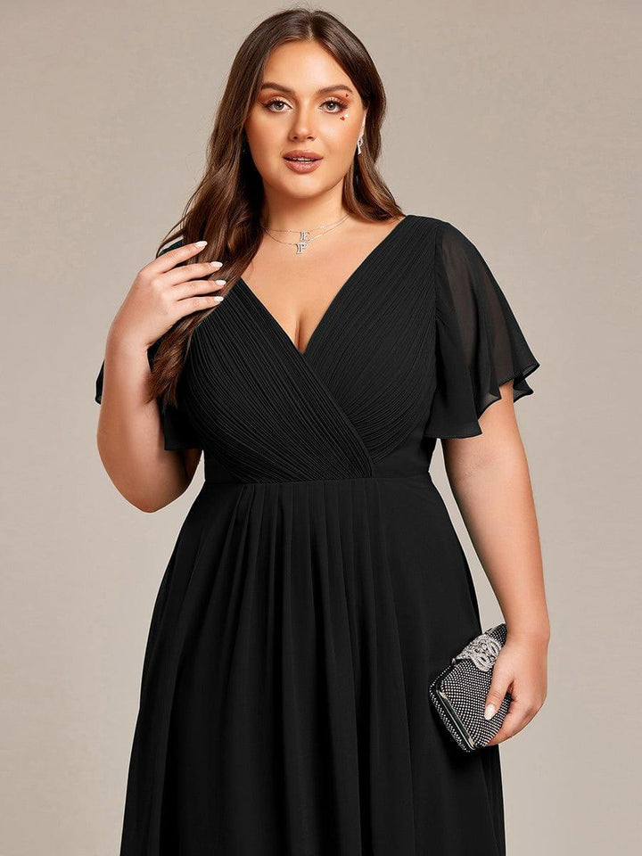 Fekete ruha felsőrésze közelről plus size modellen, redőzött V-nyak, fodros ujjak, a kezében csillogó kis táska. 28