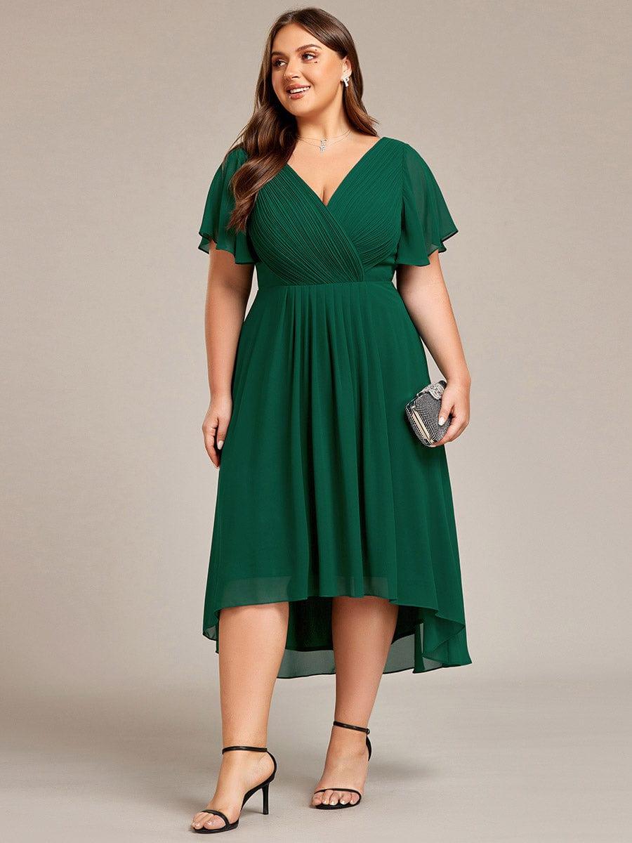 Sötétzöld ruha plus size modellen elölről, kezében csillogó kistáskával, fekete pántos magas sarkú cipőben. 40