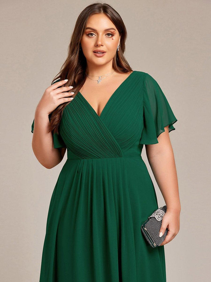 Sötétzöld ruha felsőrésze közelről plus size modellen, redőzött V-nyak, fodros ujjak, kezében csillogó kistáska. 38