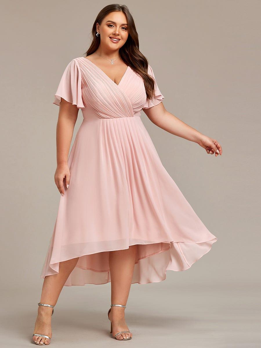 Rózsaszín ruha plus size modellen elölről, kinyújtott kézzel, a magas-alacsony szegély lágyan leng. 34