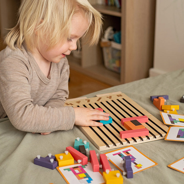 Szőke kisgyerek mosolyogva rakja ki a fa tetris puzzle-t az asztalon, körülötte mintakártyák és tüskés elemek. 4