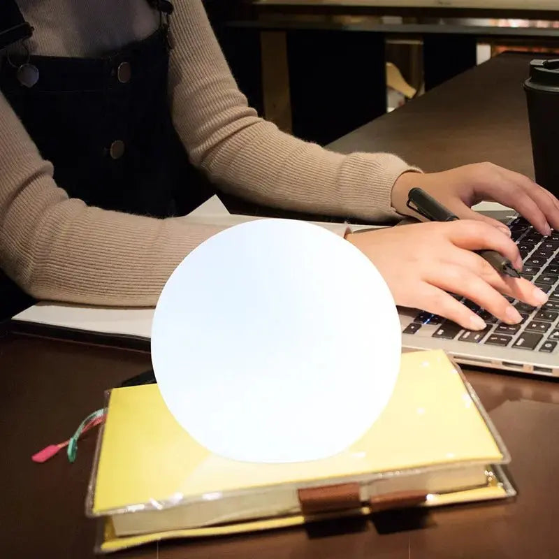 Fehéren világító gömblámpa egy jegyzetfüzeten, mögötte egy nő laptopon gépel az asztalnál. 7