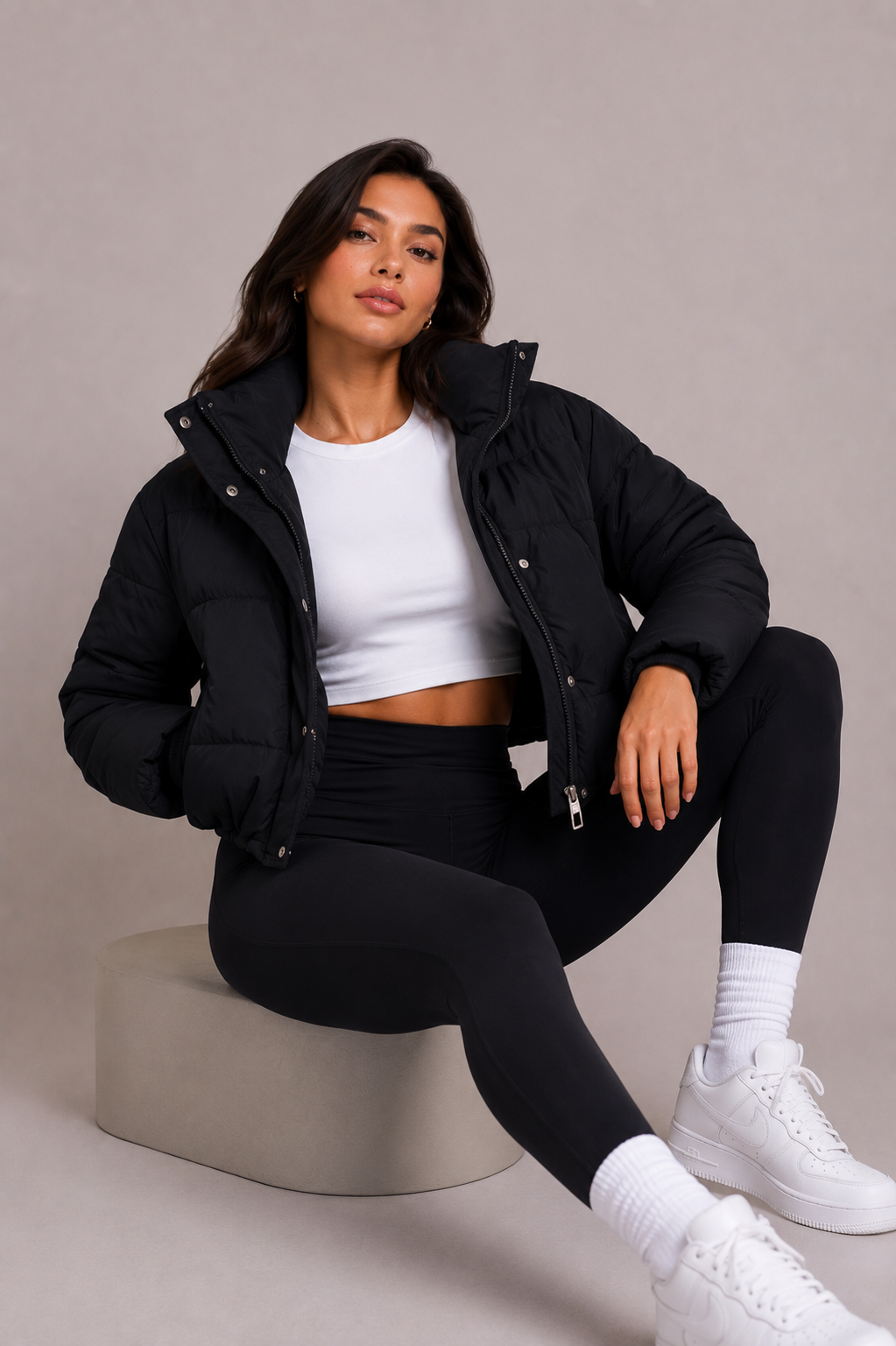 A modell egy szürke dobogón ül, nyitott fekete pufikabátban, fehér crop topban, fekete leggingsben és fehér sportcipőben. 6