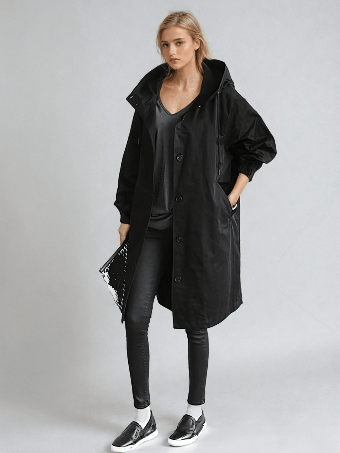 Fekete, kapucnis, gombos hosszú kabát egész alakos képen, sötét nadrággal és laposcipővel, a modell kezében csillogó pénztárca. 2