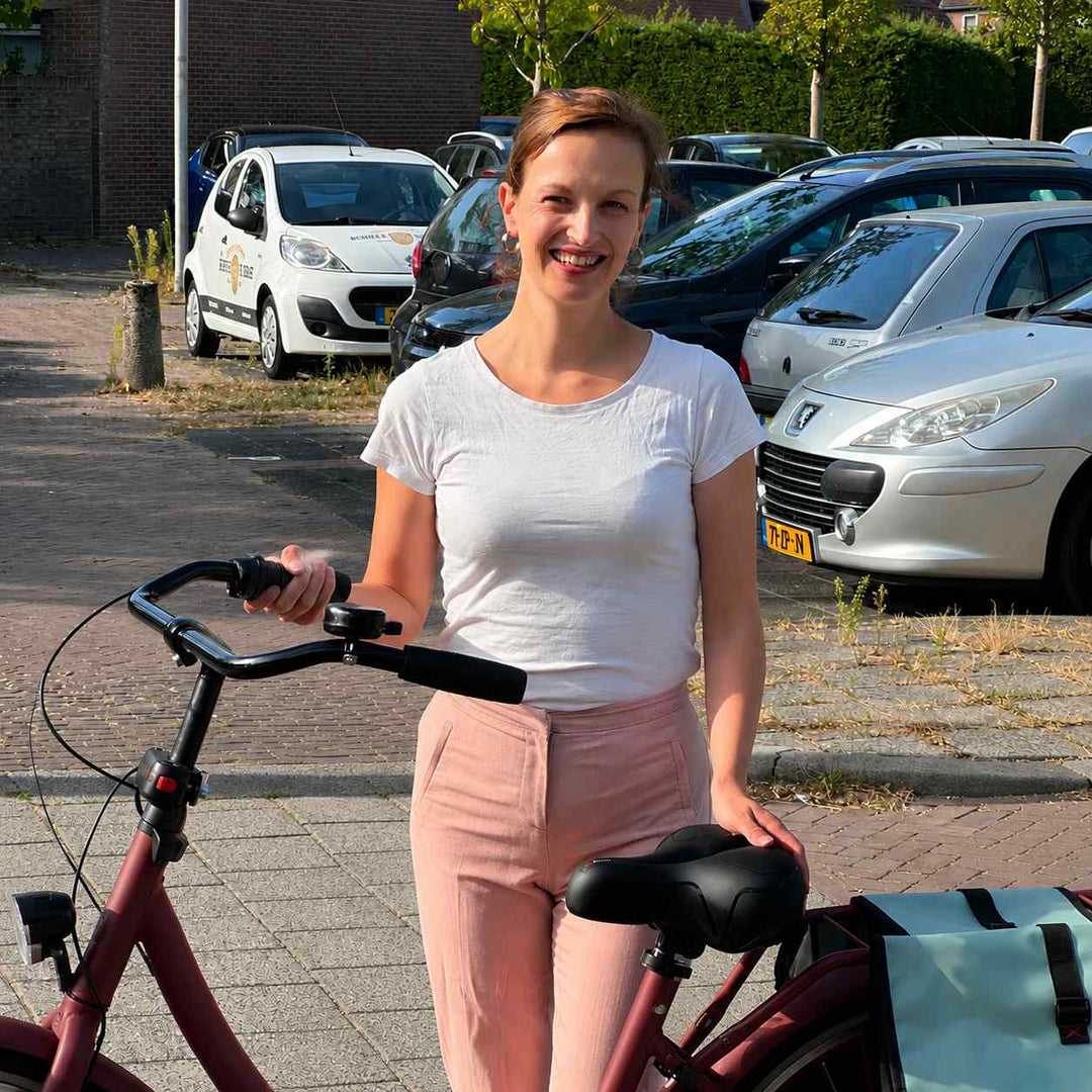 Mosolygó nő áll a piros városi kerékpár mellett, kezét a fekete nyeregen tartja, a kormányon csengő van. 7