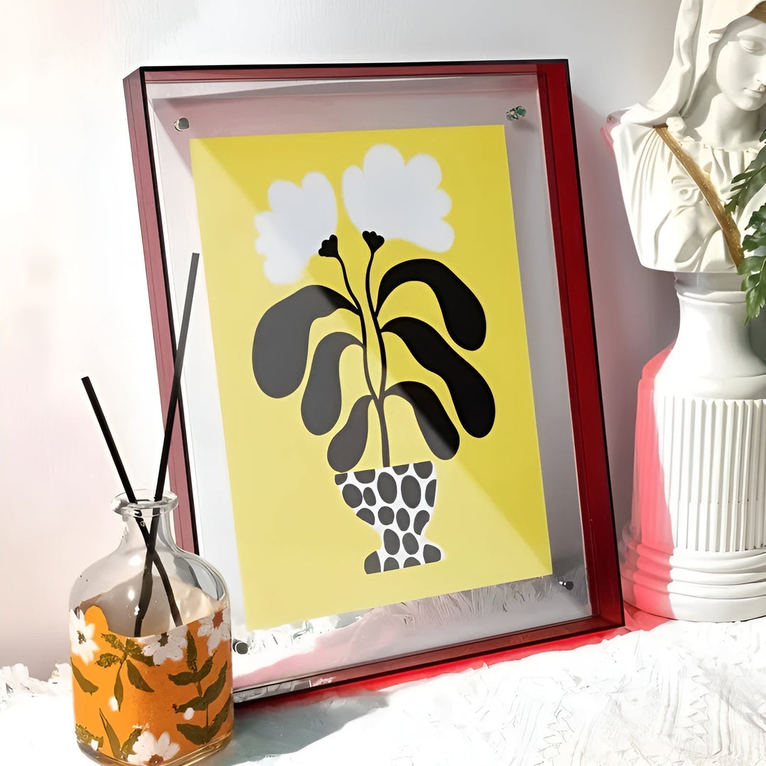 Bordó akrilkeret sárga hátterű, vázás növényes grafikával, mellette diffúzor és fehér szobor az asztalon. 15