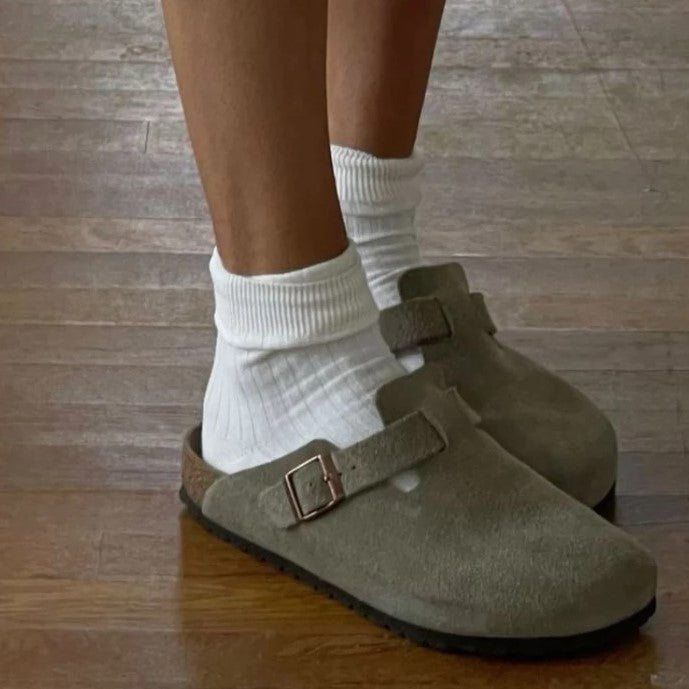 Graue Wildleder-Clogs mit Schnalle, getragen mit weißen Socken auf Holzboden.