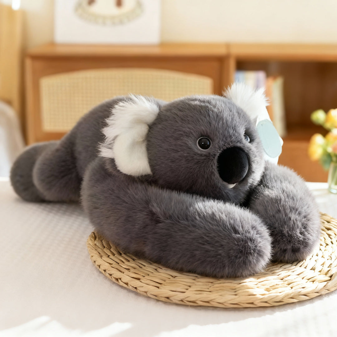 Szürke koala plüss fehér füllel és nagy fekete orral, hosszú szürke karjaival egy fonott alátéten fekve. 16